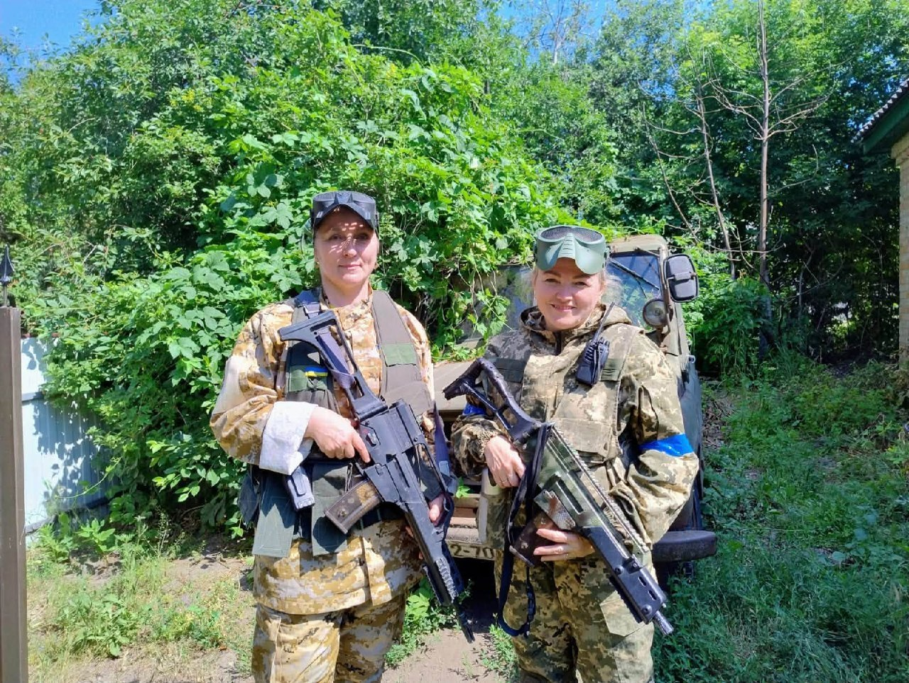 Дві жінки в камуфляжній формі та захисних окулярах стоять на тлі зелених кущів та позашляховика, тримаючи в руках ігрову зброю для лазертагу або страйкболу.