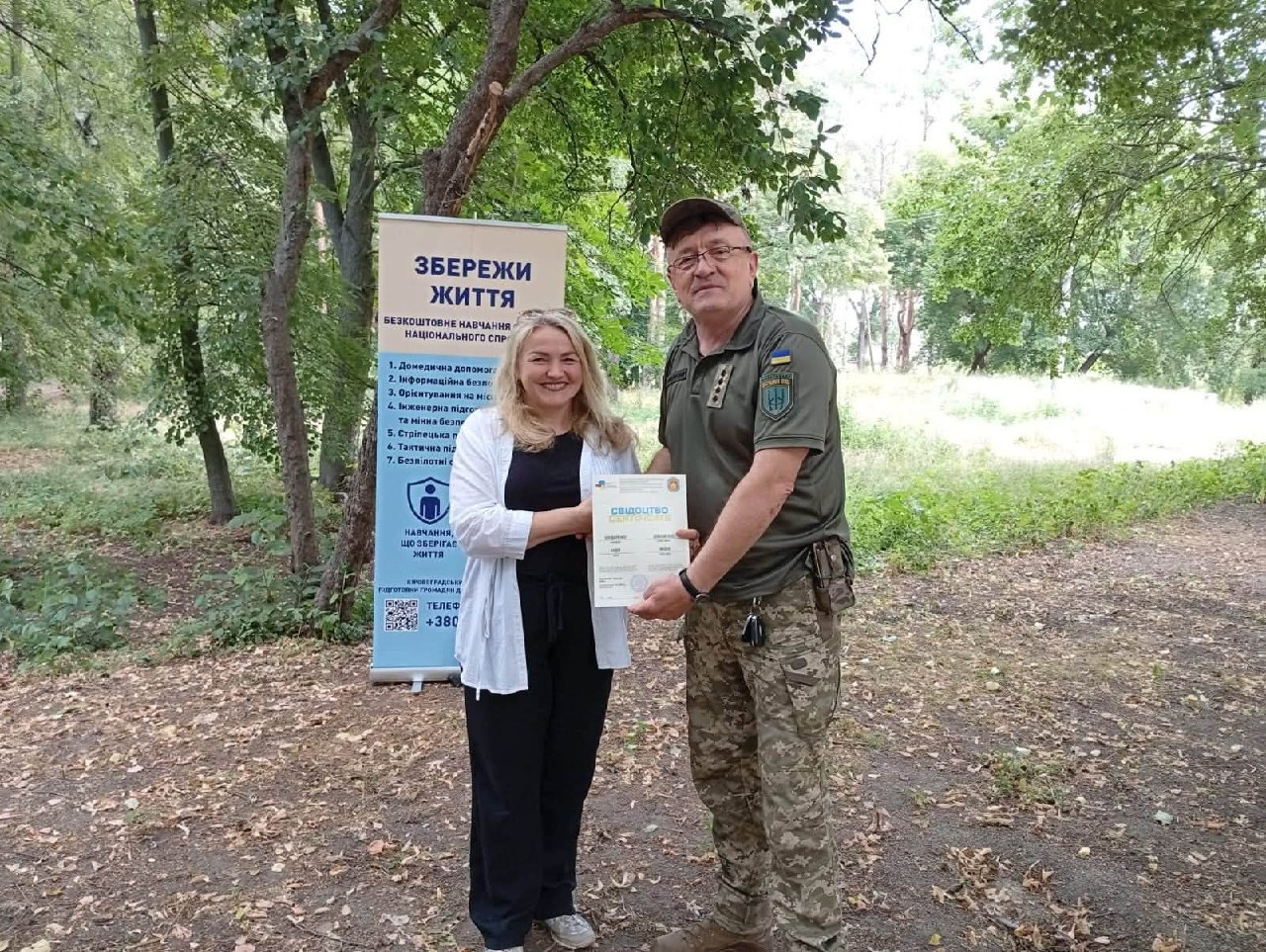 Чоловік у військовій формі вручає паперовий сертифікат жінці зі світлим волоссям, яка одягнена у світлу сорочку та чорні штани, в парку на тлі банера з написом «Збережи життя».