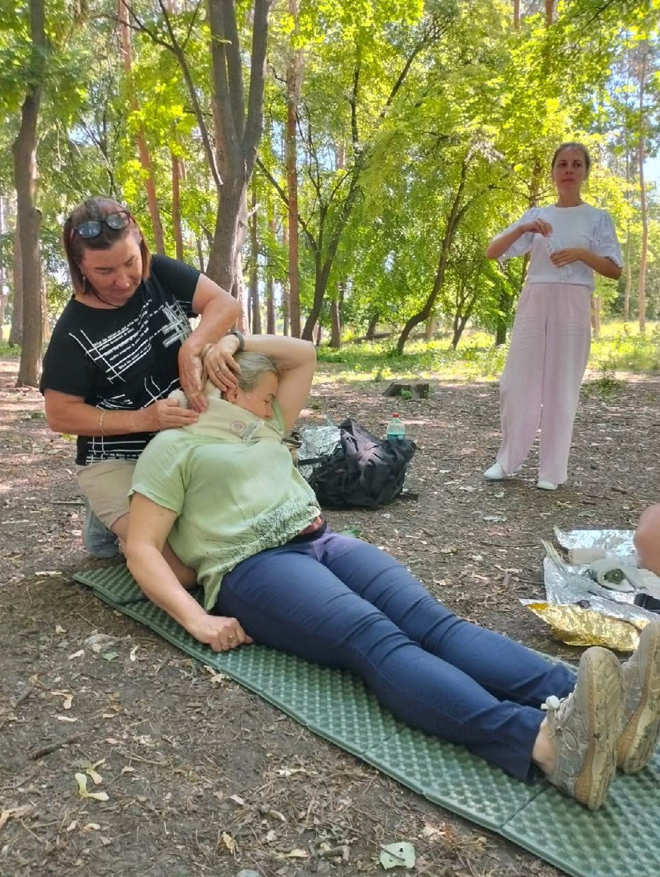 Під час практичного заняття в лісопарку жінка накладає шийний комір іншій жінці, яка напівлежить на туристичному килимку, поки на задньому плані за ними спостерігає дівчина у світлому одязі.