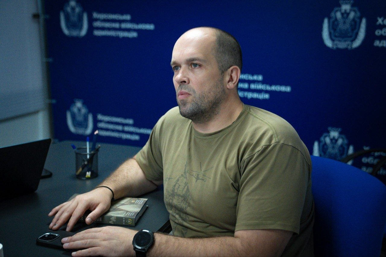 Чоловік у футболці кольору хакі сидить за столом під час зустрічі на тлі синього банера з написами та символікою. Перед ним на столі лежать книга та смартфон, а на руці одягнений смартгодинник.