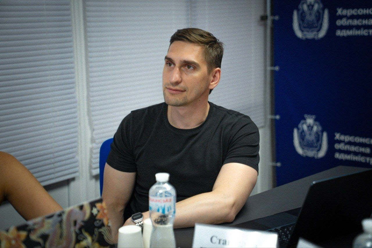 Чоловік у чорній футболці сидить за столом під час наради на тлі жалюзі та синього банера з написами й символікою. Перед ним на столі стоять пляшка з водою, паперові склянки та видно край таблички з текстом.