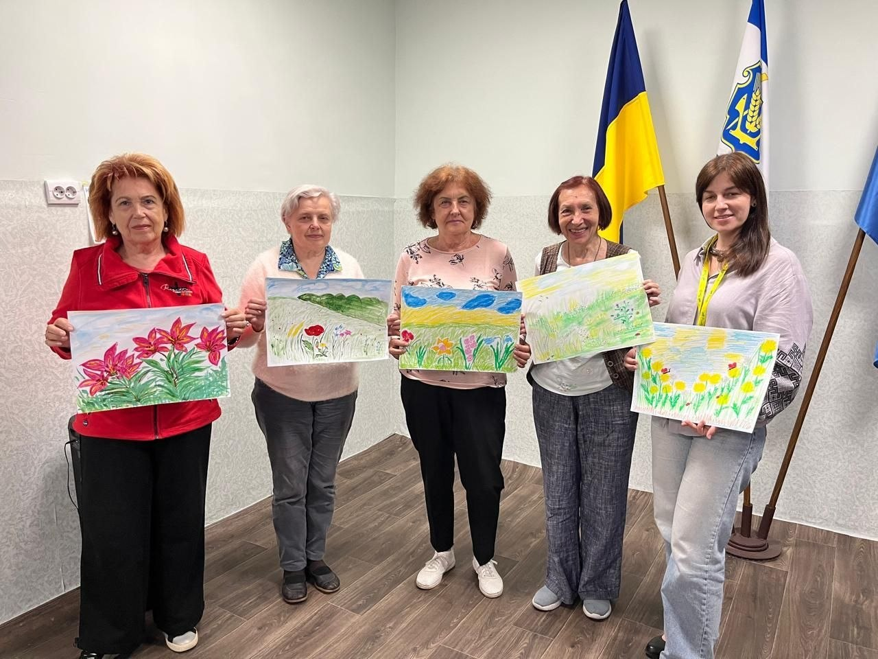 П'ять жінок стоять у ряд у кімнаті та тримають у руках власні малюнки з квітами й пейзажами на тлі прапорів.