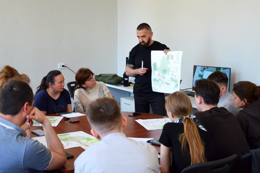 Чоловік у чорній футболці стоїть біля столу та демонструє групі людей паперову карту чи схему зелених зон. Учасники зустрічі сидять навколо круглого столу, слухають доповідача та розглядають аналогічні роздруковані матеріали. На задньому плані видно світлу стіну та монітор комп'ютера.