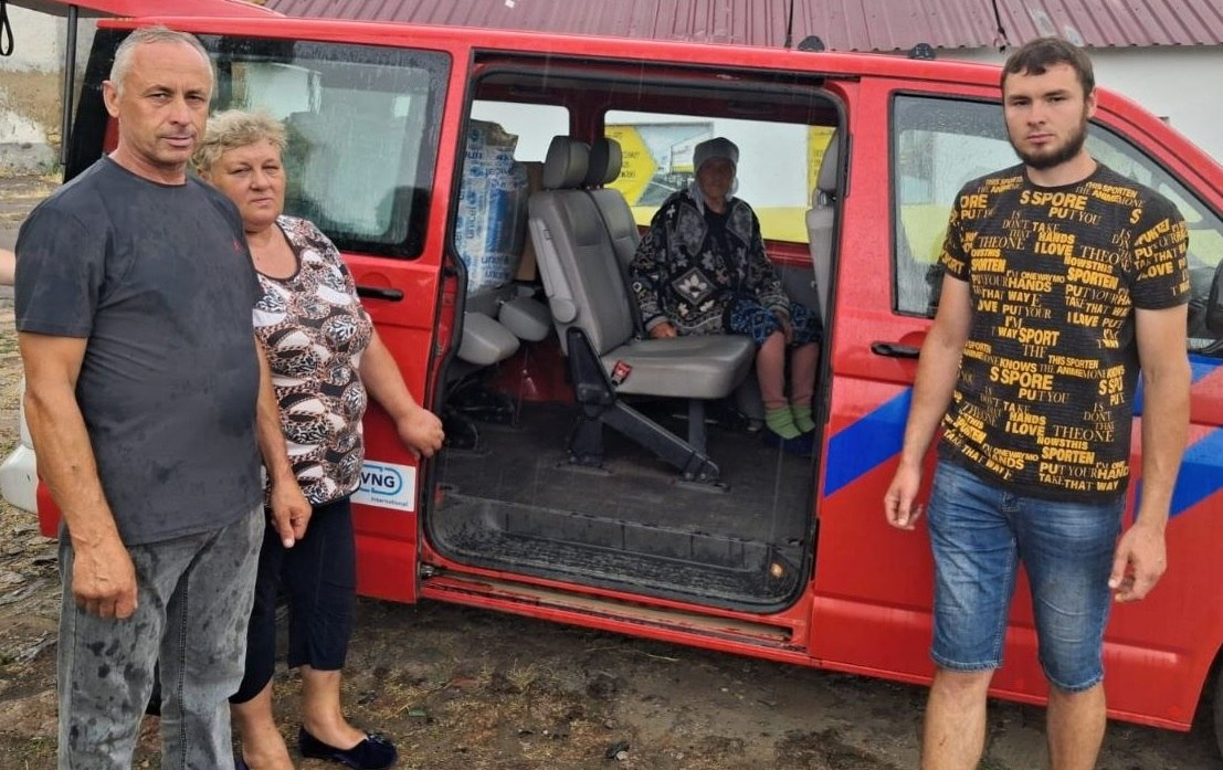 Група людей біля червоного пасажирського мікроавтобуса з відкритими дверима. Ліворуч стоять чоловік та жінка, справа — молодий чоловік. Всередині салону на сидінні перебуває літня жінка у хустці, а позаду неї видно складений вантаж або пакунки.