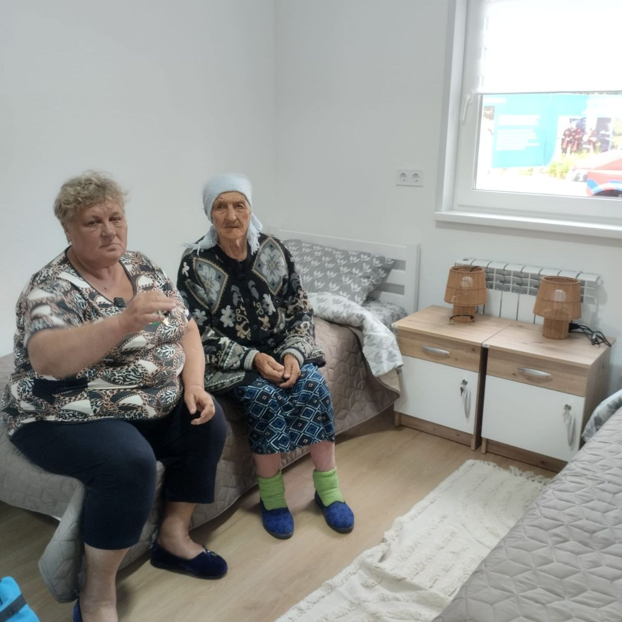 Дві жінки сидять на ліжку у світлій кімнаті. Ліворуч — жінка у візерунчастій кофті, яка тримає у руці ключі, праворуч — літня жінка у світлій хустці. Поруч стоять дві тумбочки з настільними лампами, а справа видно вікно.