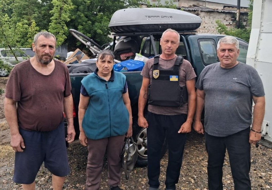 Чотири людини стоять у ряд на тлі завантаженого автомобіля-пікапа. Зліва направо: чоловік у коричневій футболці, жінка у синій безрукавці, чоловік у бронежилеті з українським прапором та сивовласий чоловік у сірій футболці. Кузов автомобіля позаду них заповнений речами, а на даху встановлено додатковий закритий багажник.