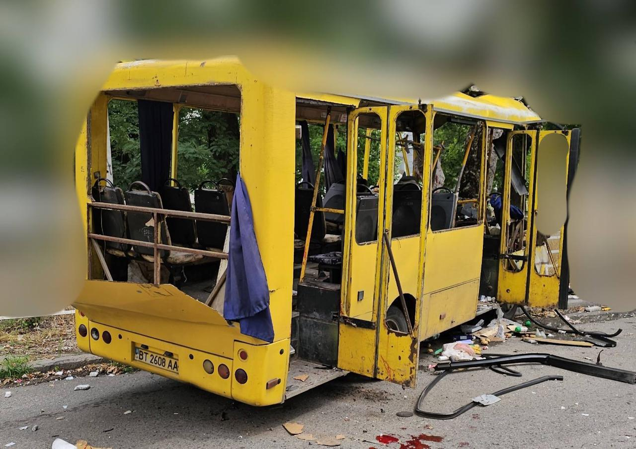 Жовтий пасажирський автобус із сильними пошкодженнями внаслідок обстрілу чи вибуху, що стоїть на дорозі. У транспортного засобу повністю вибиті вікна, зірвано задню частину кузова, деформовано дах та двері. На асфальті навколо автобуса розкидані уламки, сміття та видно сліди крові. На задньому бампері чітко видно автомобільний номерний знак.