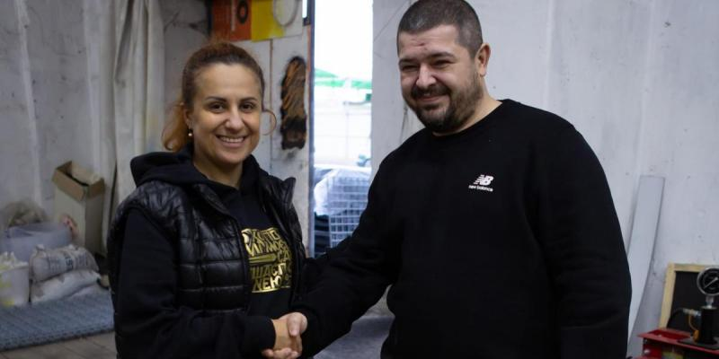 Чоловік та жінка посміхаються і тиснуть один одному руки у приміщенні майстерні чи складу. Ліворуч стоїть жінка у чорному худі, стьобаній жилетці та зі зібраним волоссям. Праворуч — бородатий чоловік у чорному світшоті бренду New Balance. На задньому плані видно світлі стіни, відкриті двері, мішки та робочі матеріали.