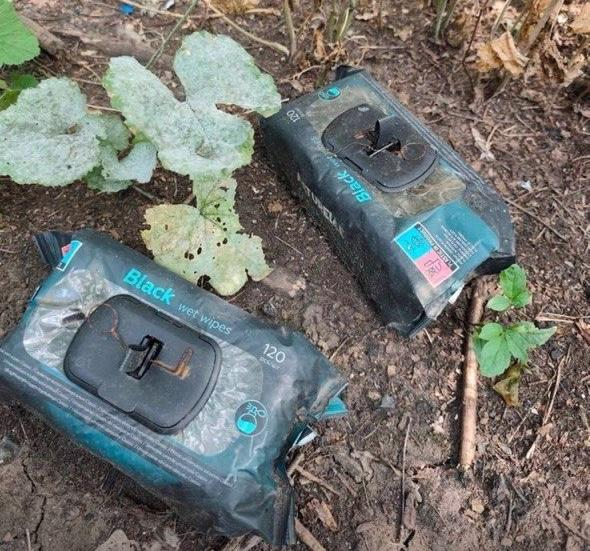 Дві великі пачки вологих серветок у темній обгортці лежать на землі серед сухого листя, гілок та зелених рослин. На пакованнях із пластиковими клапанами видно написи «Black wet wipes» та число «120». Самі пачки та земля навколо них виглядають дещо присипаними пилом або брудом.