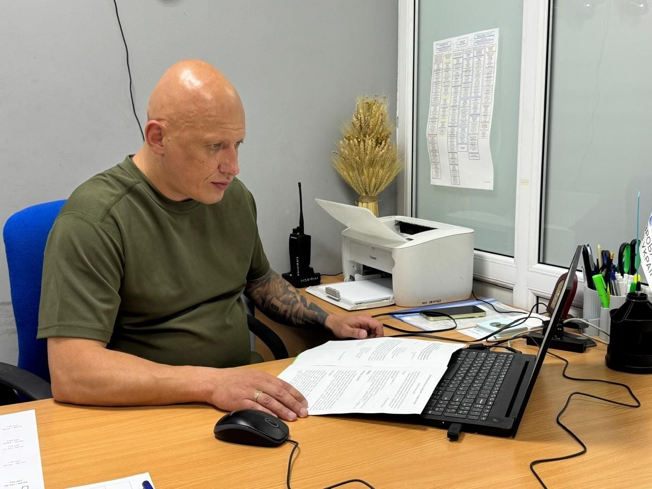 Чоловік у зеленій футболці сидит за робочим столом в офісі та дивиться в екран ноутбука. Перед ним лежать роздруковані документи, а рука знаходиться на комп'ютерній миші. На столі також стоять принтер, рація, підставка з ручками та документи на стіні й скляних дверях позаду.