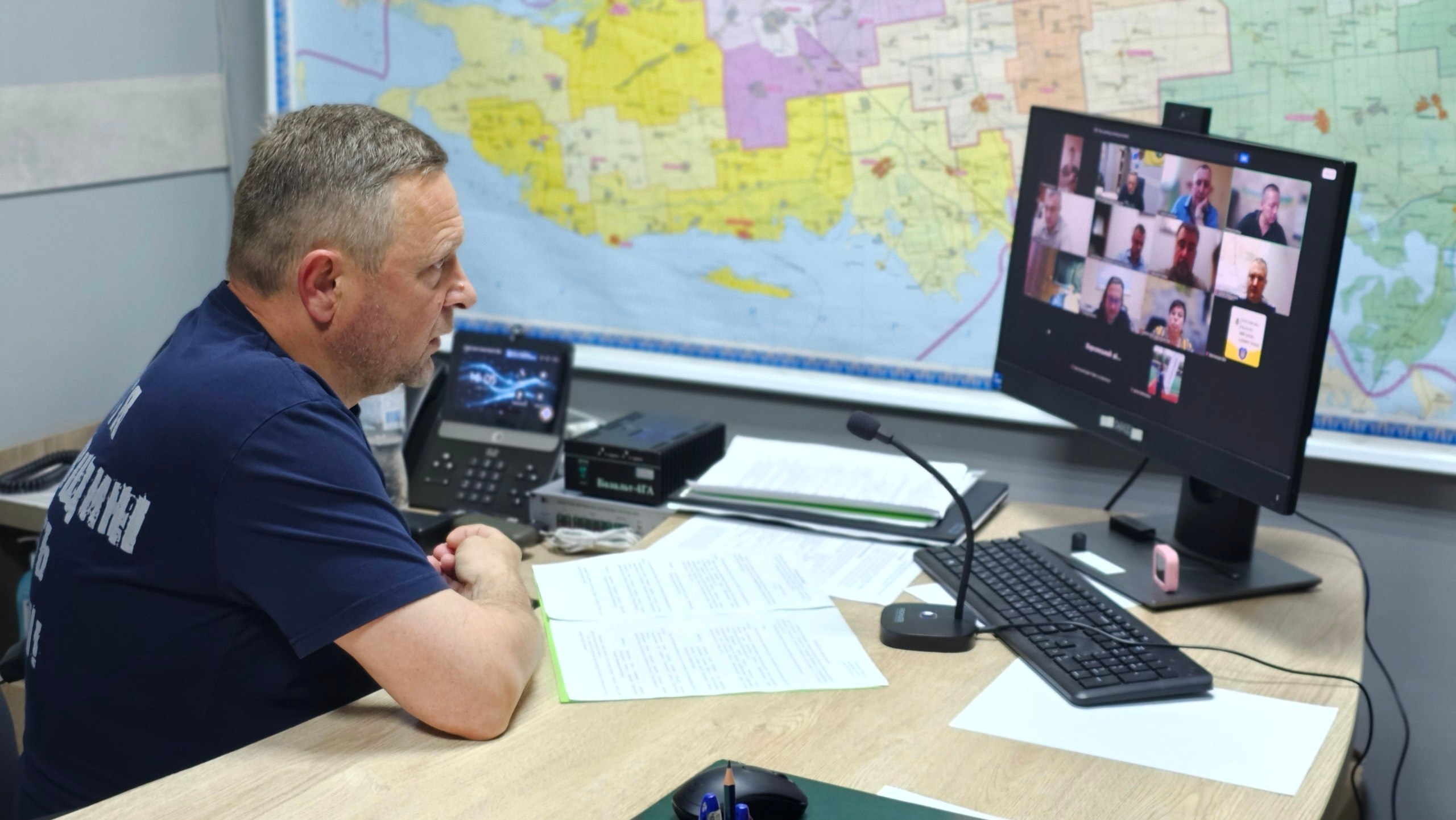 Чоловік у синій футболці сидить за робочим столом у кабінеті та дивиться на монітор комп'ютера, де триває онлайн-нарада у форматі відеоконференції з багатьма учасниками. На столі перед ним лежать документи, клавіатура, миша, мікрофон та настільний телефон. На стіні позаду монітора розміщена велика кольорова мапа.