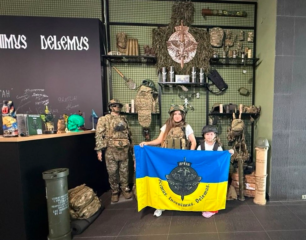 Жінка та дитина стоять у залі та тримають синьо-жовтий прапор із військовим шевроном Артилерійської розвідки та латинським написом «Scimus, Invenimus, Delemus». Поруч із ними стоїть манекен у повному військовому екіпіруванні, а позаду розміщено тематичний стенд із військовим спорядженням, камуфляжем, рюкзаками та гільзами.