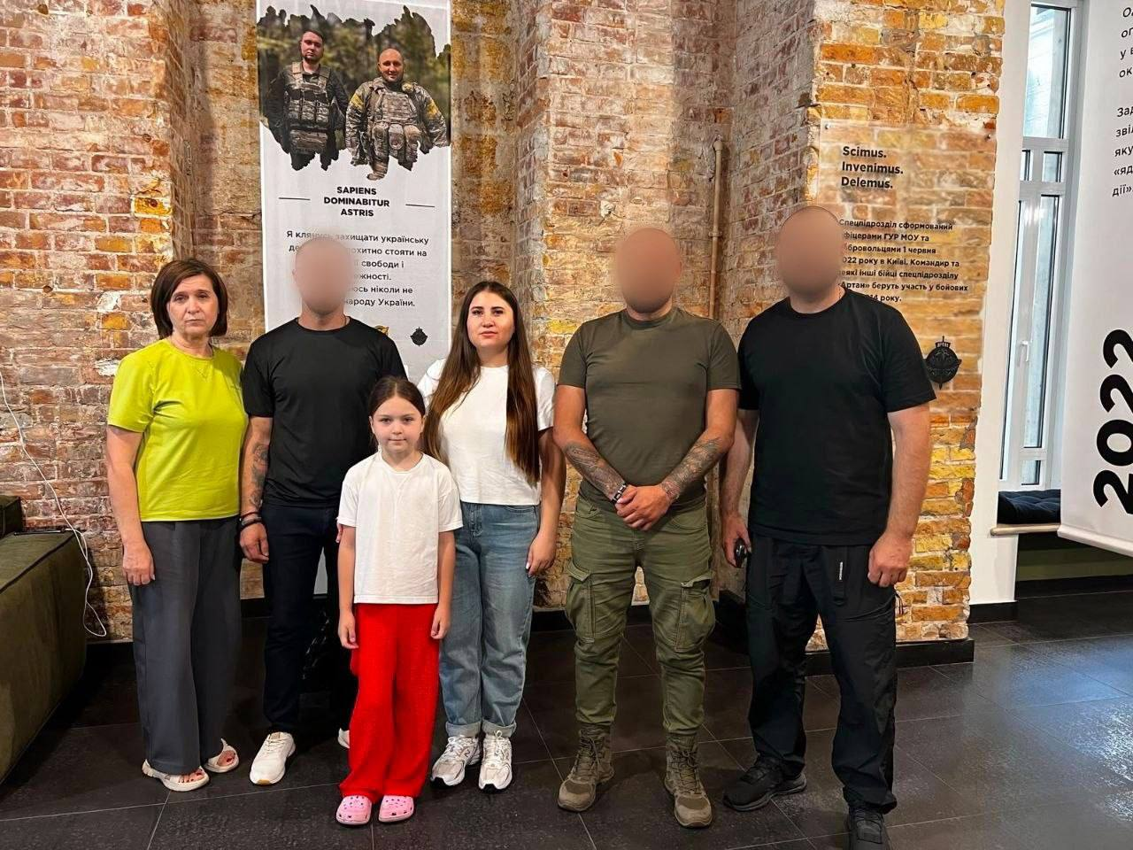 Група людей з шести осіб, серед яких дві жінки, дитина та троє чоловіків у мілітарі одязі, стоять у приміщенні з цегляними стінами. Обличчя трьох чоловіків приховані (розмиті), а обличчя жінок та дитини залишаються відкритими. На задньому плані розміщено інформаційні стенди з військовою символікою.