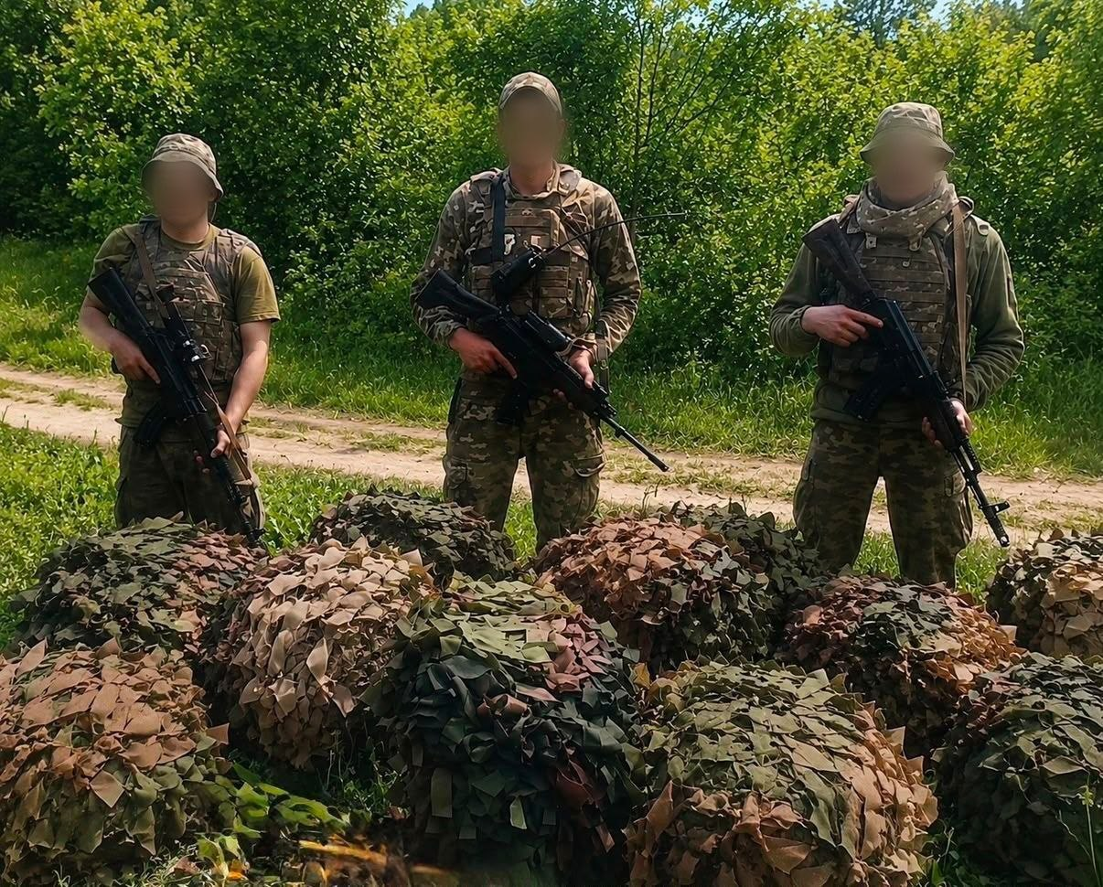 Троє військових у камуфляжній формі та захисному спорядженні стоять зі зброєю в руках на тлі зелених кущів і дерев. Обличчя всіх бійців приховані (розмиті). Перед ними на землі складені згорнуті маскувальні сітки природних зелених та коричневих відтінків.