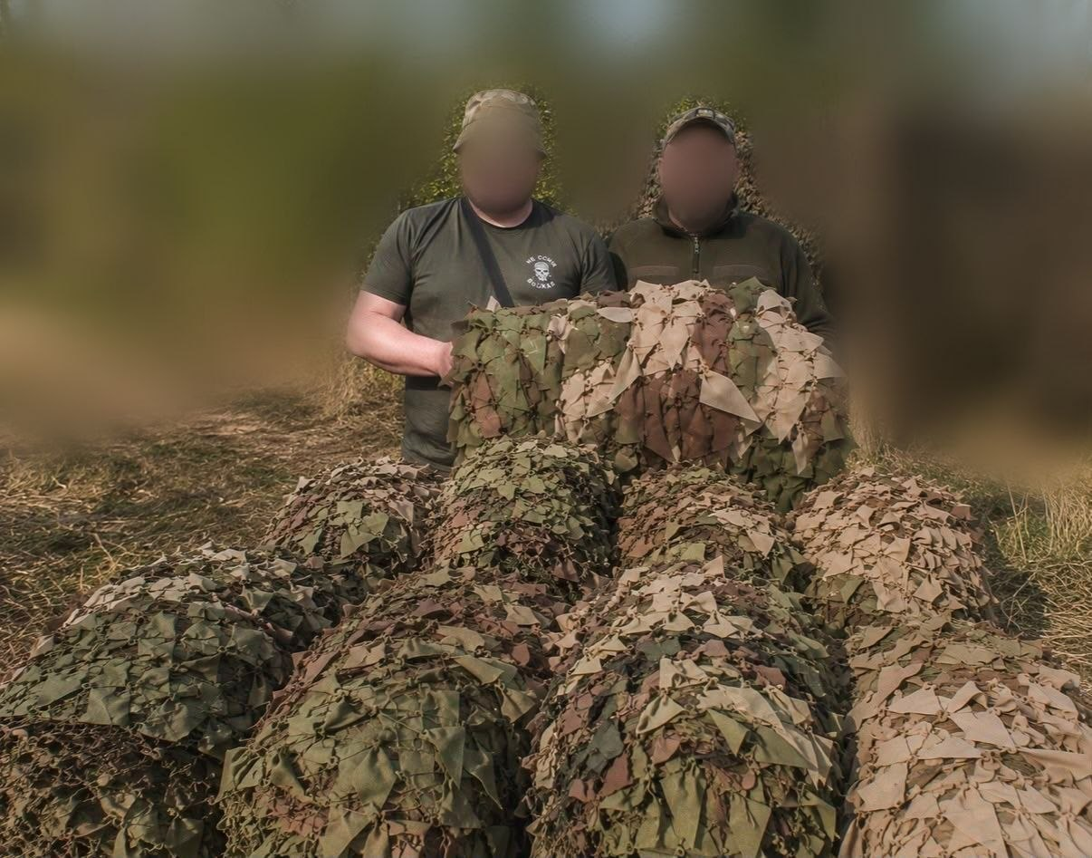 Двоє чоловіків у мілітарі одязі стоять просто неба на тлі природи, тримаючи великий сувій маскувальної сітки. Перед ними на землі складено ще кілька згорнутих маскувальних сіток зелених та коричневих відтінків. Обличчя обох чоловіків та задній план приховані (розмиті).