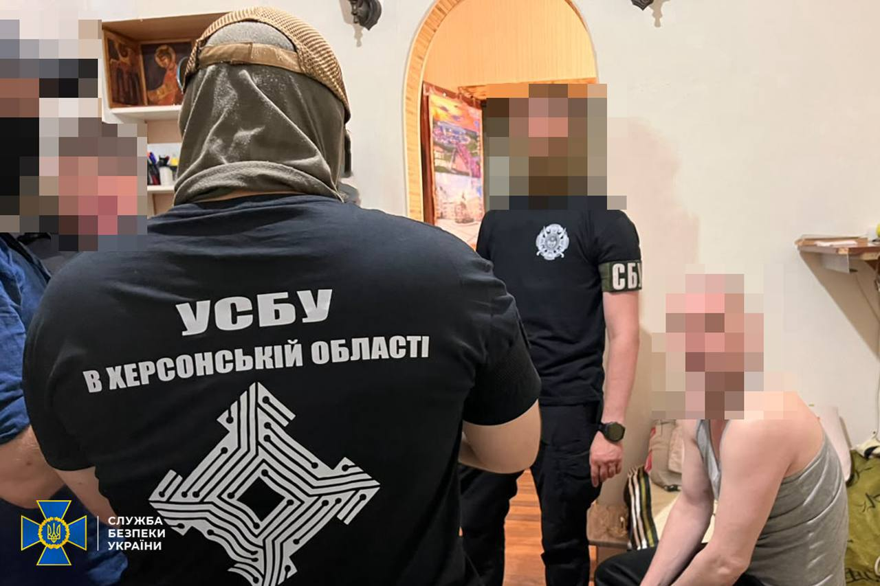 Співробітник СБУ у чорній футболці з написом «УСБУ В ХЕРСОНСЬКІЙ ОБЛАСТІ» та емблемою стоїть спиною до камери в приміщенні під час проведення слідчих дій. На задньому плані видно інших оперативників та цивільного чоловіка в майці. Обличчя всіх присутніх приховані (пікселізовані).