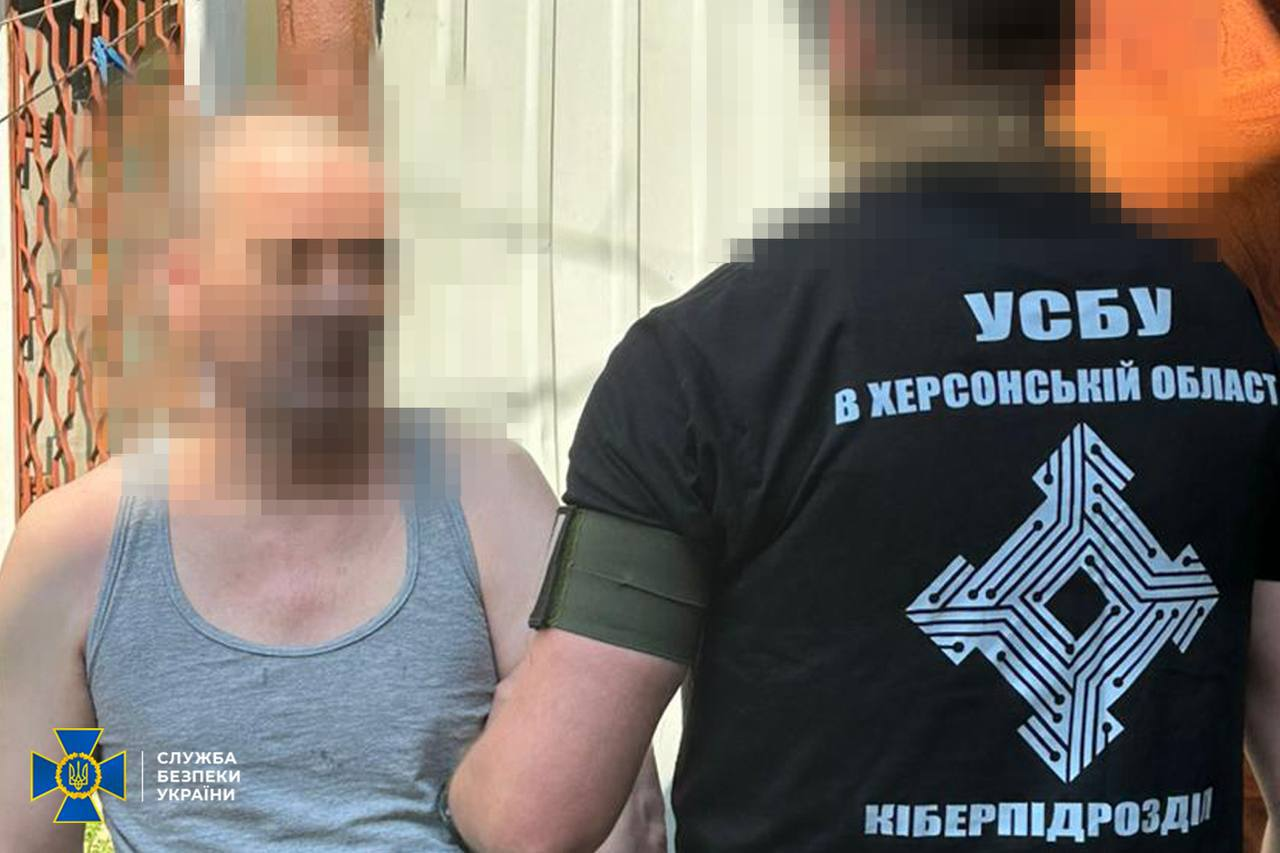 Співробітник СБУ у чорній футболці з написом «УСБУ В ХЕРСОНСЬКІЙ ОБЛАСТІ» та «КІБЕРПІДРОЗДІЛ» стоїть спиною до камери поруч із затриманим цивільним чоловіком у сірій майці. Обличчя обох чоловіків та задній план приховані (пікселізовані).