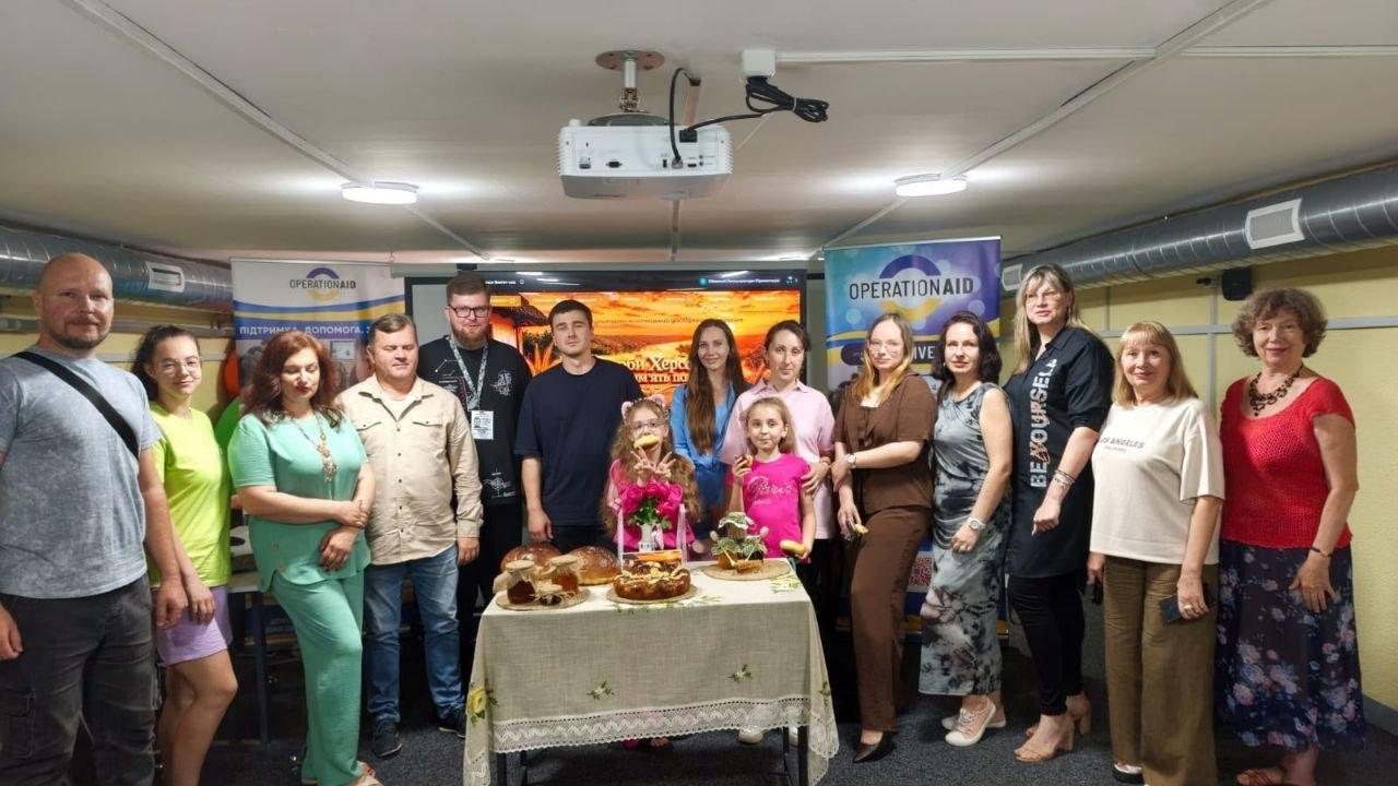 Групова фотографія понад десятка людей у приміщенні біля столу зі святковою випічкою та десертами. На задньому плані видно великий екран та два інформаційні банери благодійної організації «OPERATION AID».