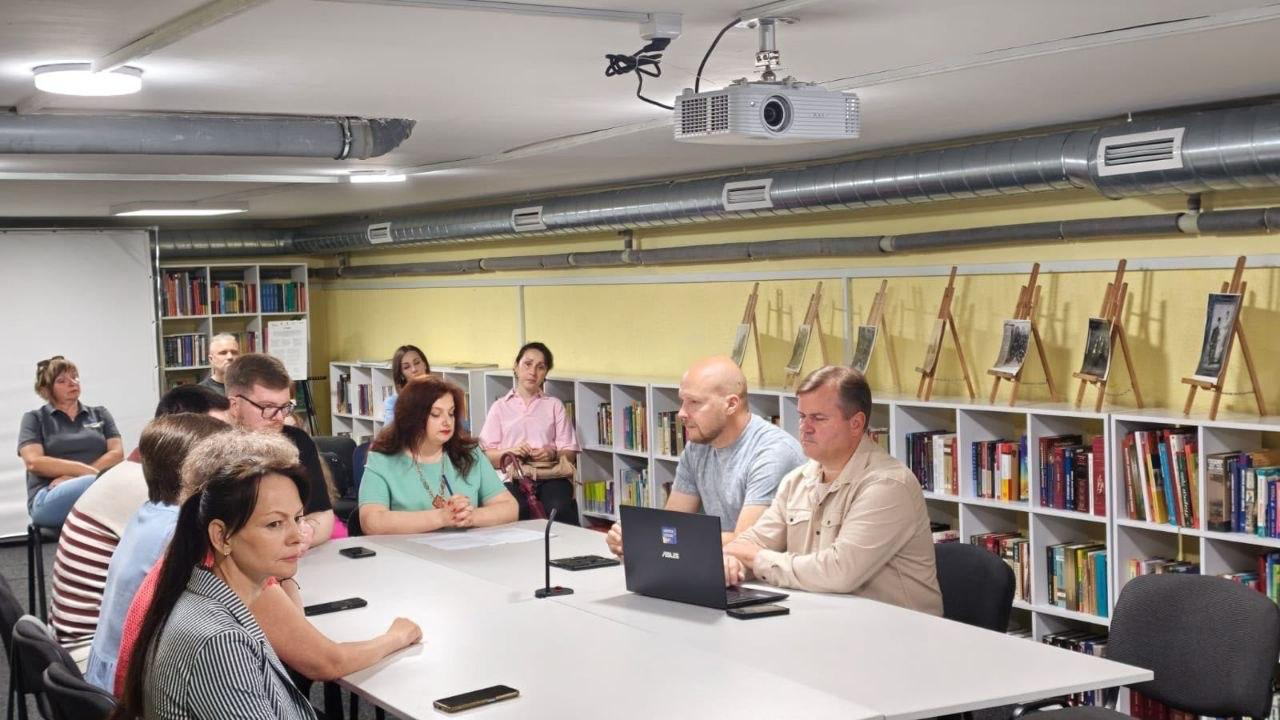 Група людей сидить за великим столом у приміщенні бібліотеки під час офіційної зустрічі або презентації. На столі перед ними стоїть відкритий ноутбук та настільний мікрофон, а на задньому плані видно книжкові полиці та невеликі мольберти з фотографіями.