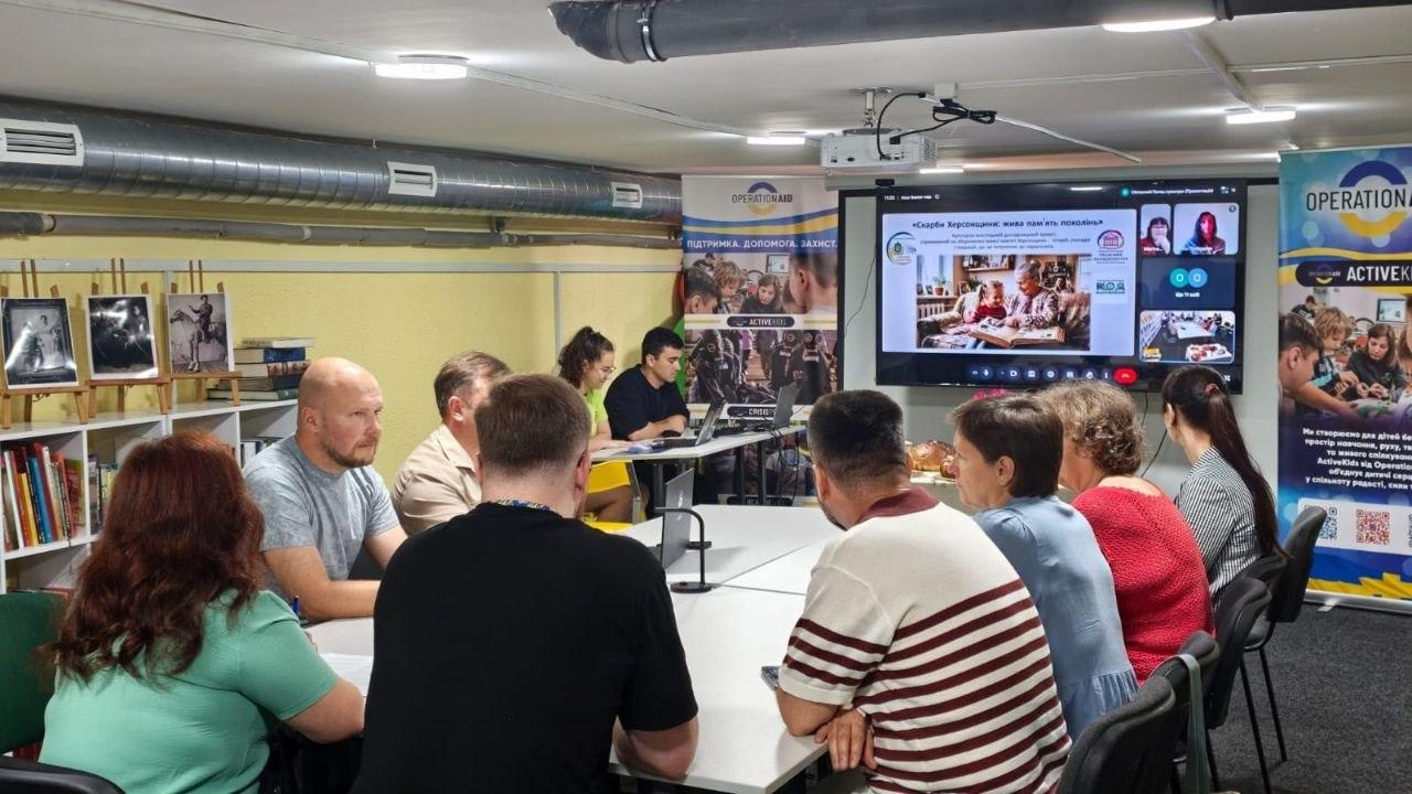 Учасники зустрічі сидять за великим столом у приміщенні бібліотеки й дивляться на великий настінний екран, де транслюється презентація та вікна відеозв'язку онлайн-наради. На задньому плані розміщено книжкові полиці з мольбертами та інформаційні банери благодійної організації «OPERATION AID».
