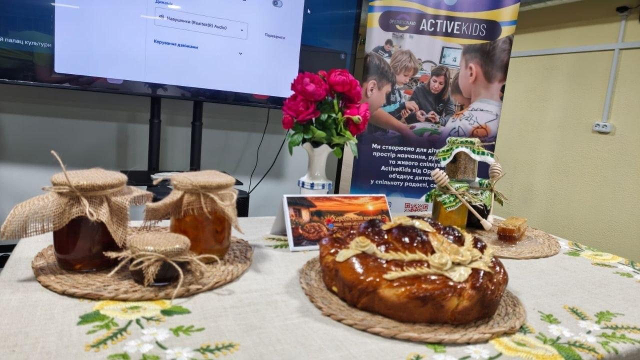 Стіл зі святковим частуванням, на якому стоїть коровай із декоративними квітами з тіста, баночки з медом та варенням, прикрашені мішковиною, а також ваза з яскраво-рожевими квітами. На задньому плані видно частину великого екрана та інформаційний банер дитячого проєкту «ACTIVEKIDS» від «OPERATION AID».