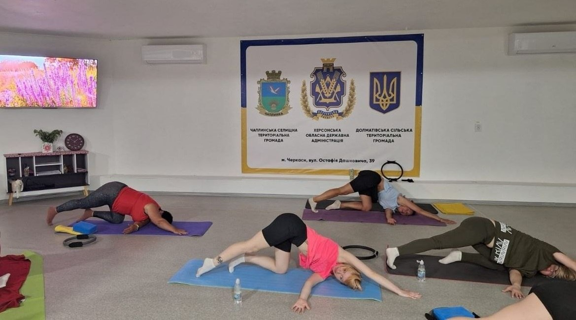 Жінки займаються йогою на килимках у залі, виконуючи вправи на розтяжку. На стіні позаду них розміщено великий банер із гербами Чаплинської, Долматівської громад, Херсонської ОДА та прапором України.