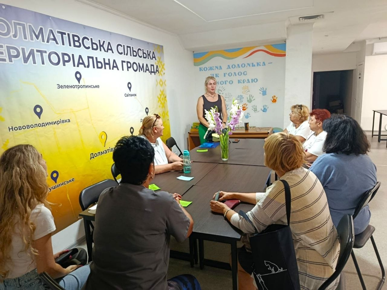 Група жінок сидить за великим столом у приміщенні під час робочої зустрічі чи обговорення. Одна з учасниць стоїть наприкінці столу та виступає перед присутніми. Ліворуч на стіні розміщено великий жовто-блакитний банер із написом «Долматівська сільська територіальна громада» та назвами населених пунктів.
