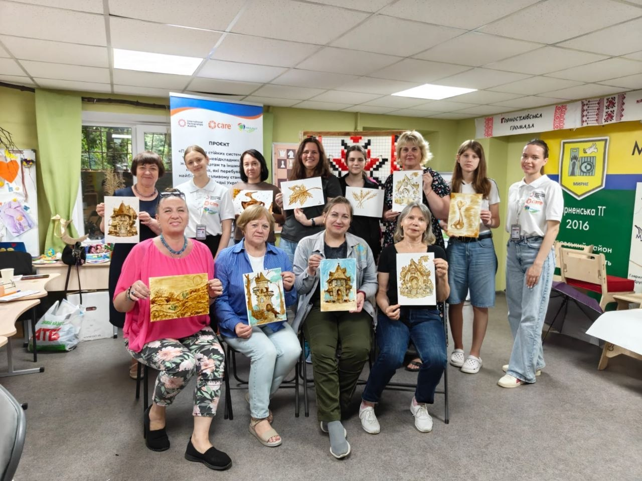 Група жінок та дівчат позує для спільного фото в приміщенні, тримаючи в руках власні художні роботи з малюнками казкових будиночків та природних мотивів. На задньому плані розміщено інформаційні банери з логотипами «Care» та символікою Горностаївської й Мирненської громад.