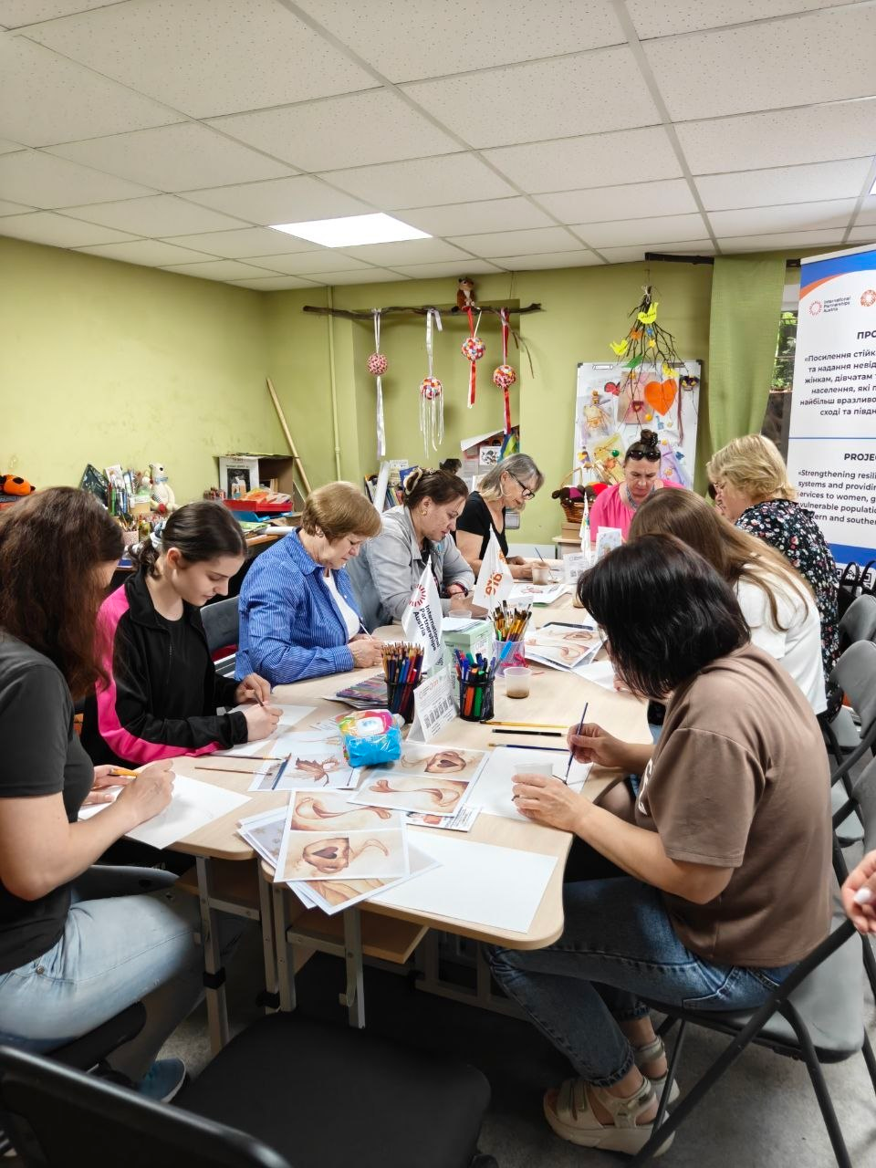 Група жінок та дівчат сидить за зсунутими столами у приміщенні під час творчого майстер-класу з малювання. Вони зосереджено працюють пензлями та олівцями, використовуючи роздруковані зразки малюнків. На столах стоять склянки з пензликами, набори олівців та маленькі настільні прапорці. Праворуч на задньому плані видно частину інформаційного банера.