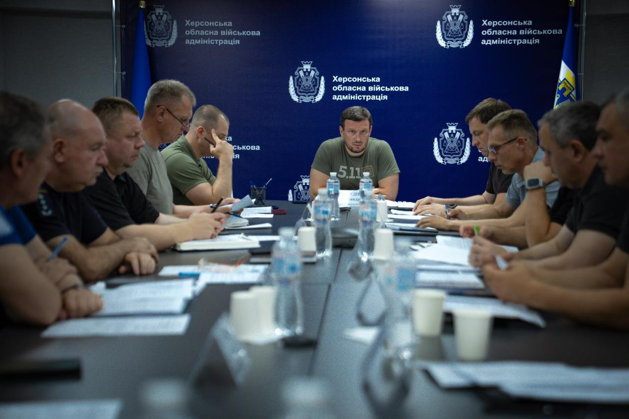 Робоча нарада за великим столом під керівництвом чоловіка у футболці кольору хакі, який сидить на чолі. По обидва боки столу розмістилися інші учасники заходу з документами та блокнотами. На задньому плані встановлено синій брендований банер з офіційною символікою та написами.