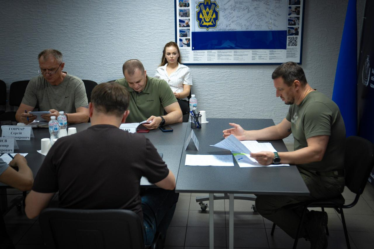 Робоча нарада в кабінеті за великим кутовим столом. Чоловік праворуч у футболці кольору хакі переглядає папери, а інші учасники зустрічі вивчають документи та роблять записи. На стіні позаду них висить великий синій плакат з офіційною символікою та підписами.