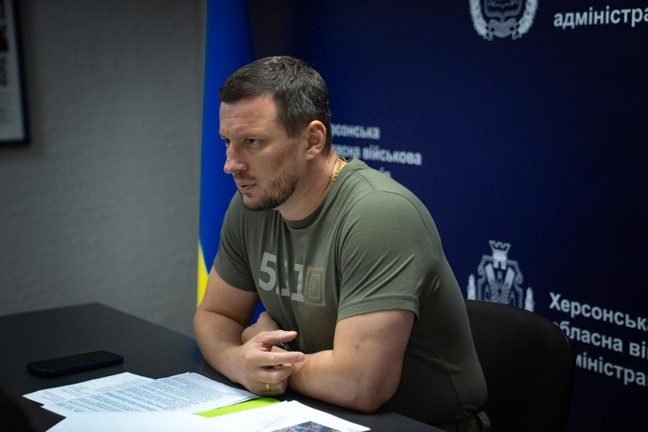 Чоловік у футболці кольору хакі сидить за столом під час наради, склавши руки перед собою, та зосереджено слухає або дивиться вбік. Перед ним на столі лежать документи та мобільний телефон. На задньому плані видно частину синьо-жовтого прапора та темно-синій брендований банер з офіційною символікою та написами.