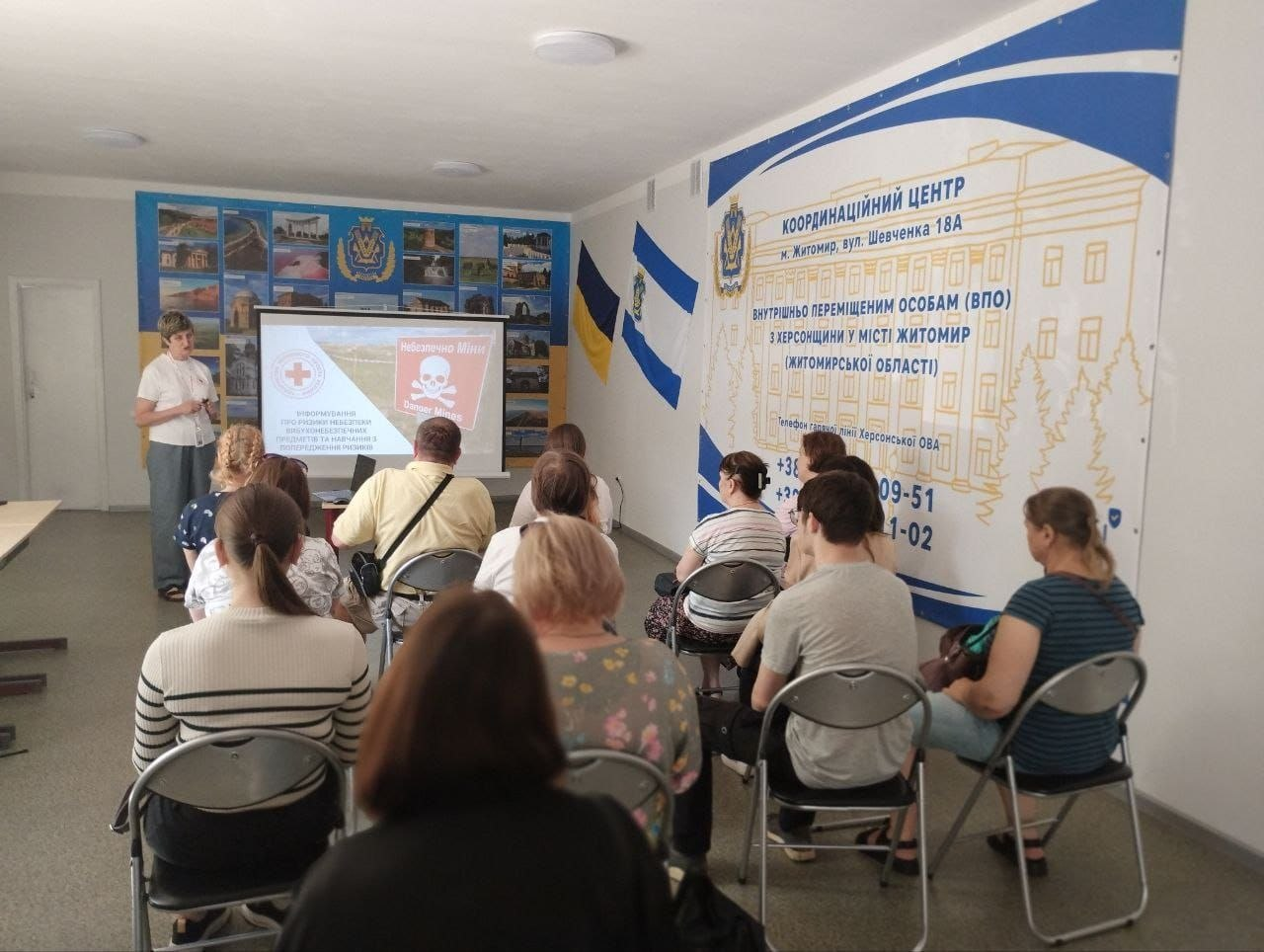 Жінка проводить лекцію з мінної безпеки для групи людей, які сидять на стільцях у приміщенні хабу. На екрані проєктора демонструється слайд із попереджувальним знаком «Небезпечно міни!». Праворуч на стіні розміщено великий банер Координаційного центру для внутрішньо переміщених осіб з Херсонщини.