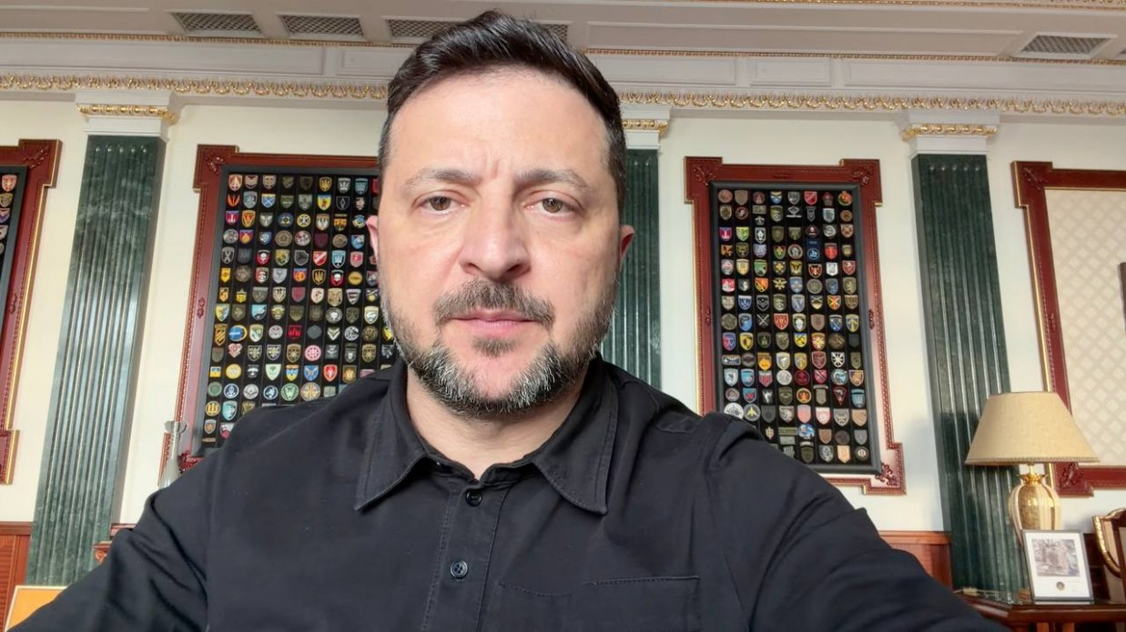Чоловік із короткою борідкою у чорній сорочці записав крупним планом відеозвернення в офіційному кабінеті. Позаду нього на стіні між колонами висять два великі прямокутні стенди у дерев'яних рамках, заповнені численними військовими шевронами та емблемами.