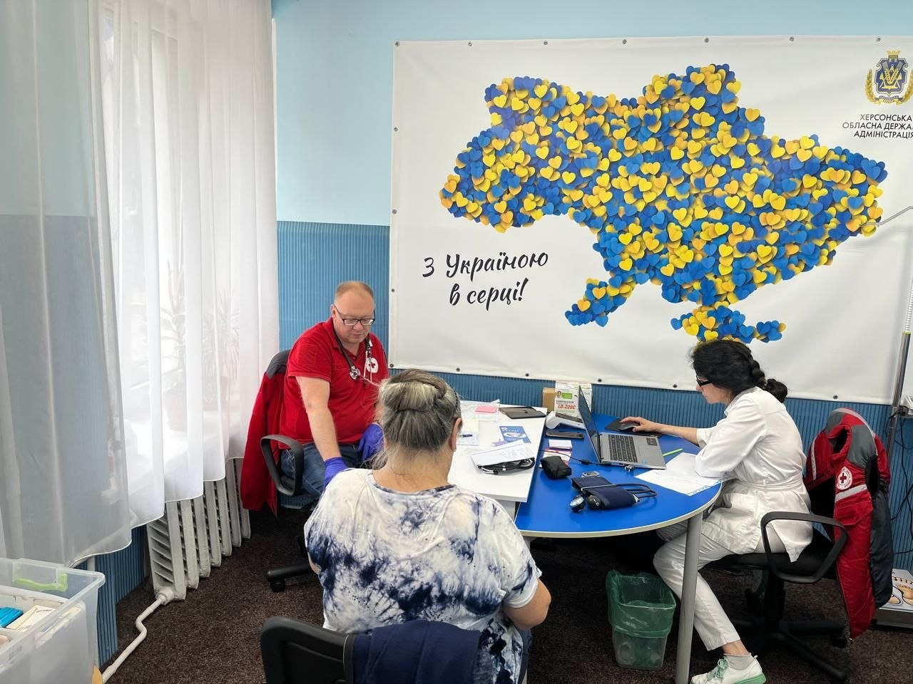 Медичні працівники приймають пацієнтку в кабінеті. На стіні розміщено банер із мапою України з жовто-блакитних сердець, написом «З Україною в серці!» та логотипом Херсонської обласної державної адміністрації.