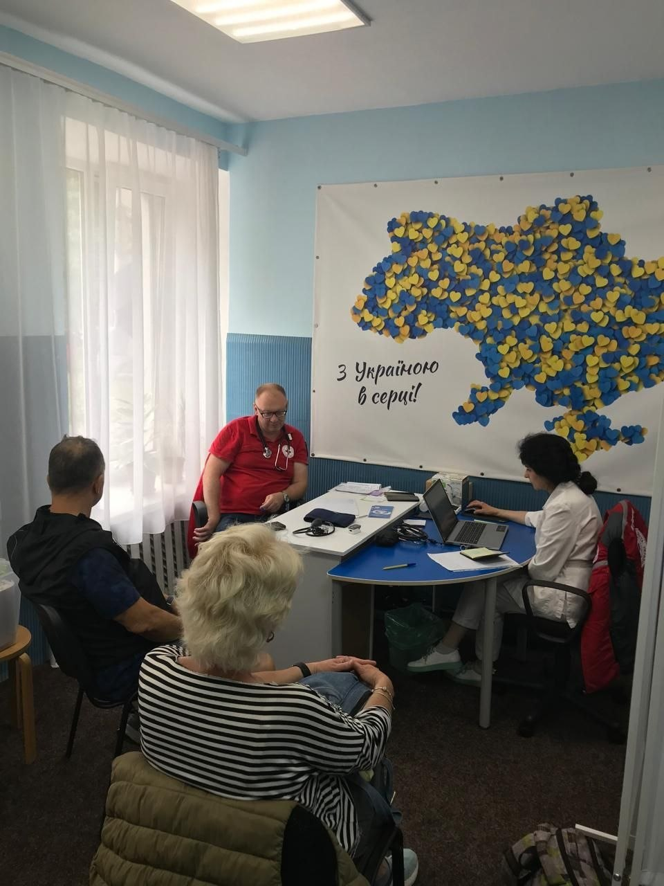 Медичні працівники консультують двох пацієнтів у кабінеті. На стіні позаду них висить банер із контуром мапи України з жовто-блакитних сердець та написом «З Україною в серці!».