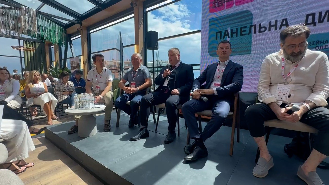 Панельна дискусія в межах офіційного форуму у світлому залі з панорамними вікнами та скляною стелею. П'ять спікерів-чоловіків сидять у президії на низькому подіумі на тлі великого екрана, а ліворуч на стільцях розмістилися слухачі заходу.