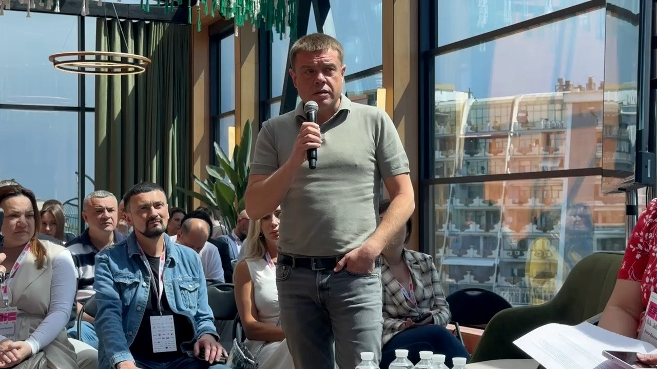 Чоловік виступає з мікрофоном під час офіційного заходу у світлій залі з панорамними вікнами. Позаду та ліворуч від нього на стільцях сидять слухачі, які уважно слухають промову.