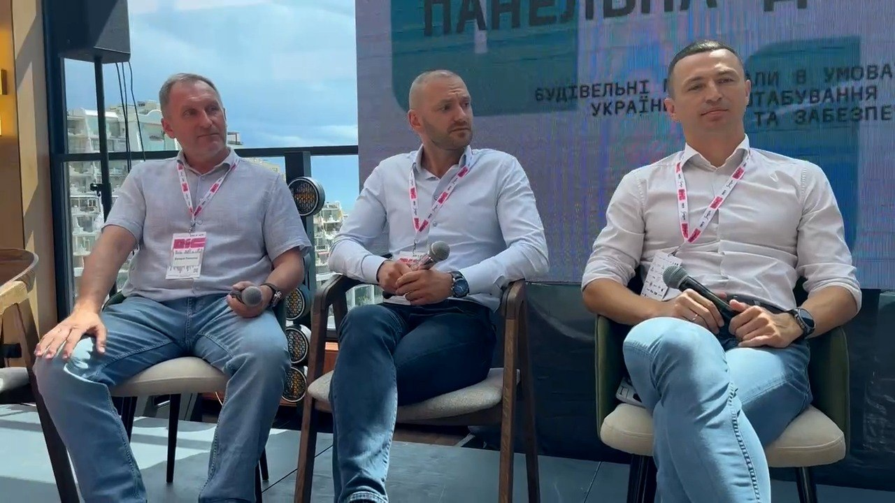 Троє спікерів-чоловіків сидять у кріслах на президії під час офіційного форуму. Вони одягнені в сорочки та джинси, мають брендовані бейджі на шиї та тримають у руках мікрофони на тлі великого екрана.