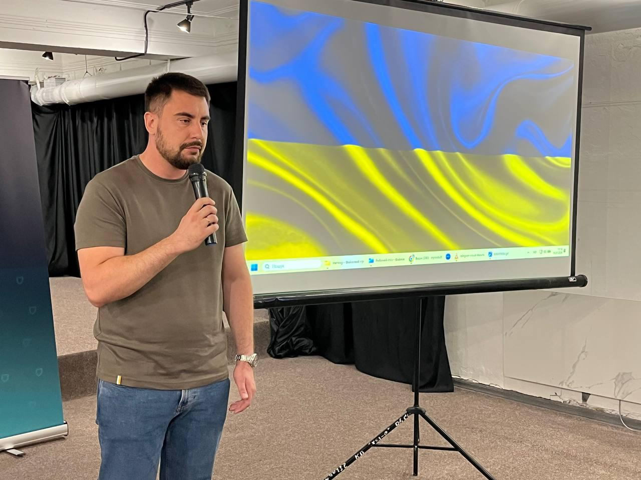 Чоловік із мікрофоном у руці виступає перед проєкторним екраном, на якому відтворюється зображення державного прапора України. Подія проходить у приміщенні з темним завісом на задньому плані, що відповідає атмосфері заходу проєкту «Пліч-о-пліч: згуртовані громади».
