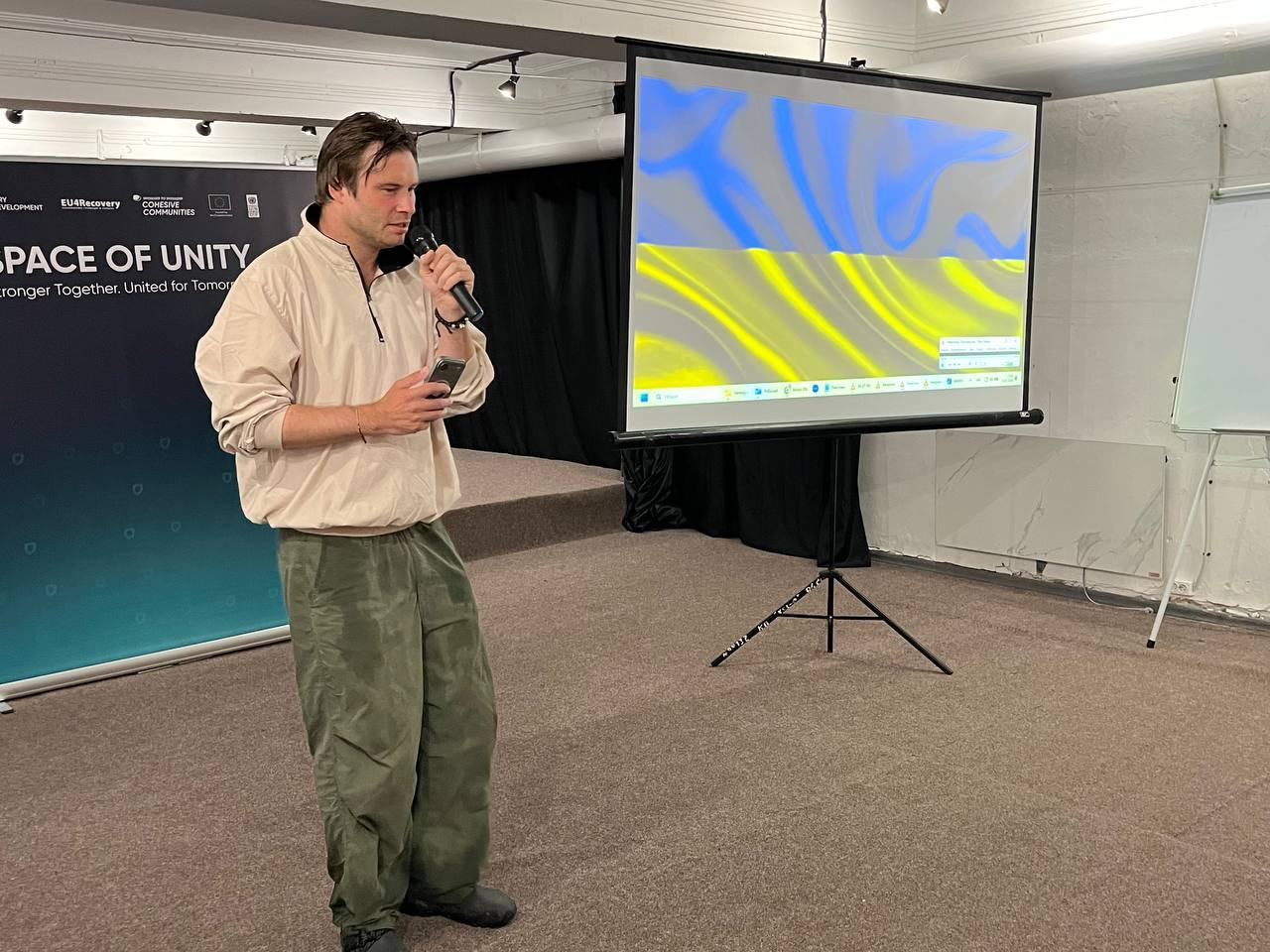 Чоловік виступає з мікрофоном у руці під час заходу проєкту «Пліч-о-пліч: згуртовані громади». Він стоїть у залі-укритті біля великого банера з написом «SPACE OF UNITY» та проєкторного екрана, на якому відображається прапор України.