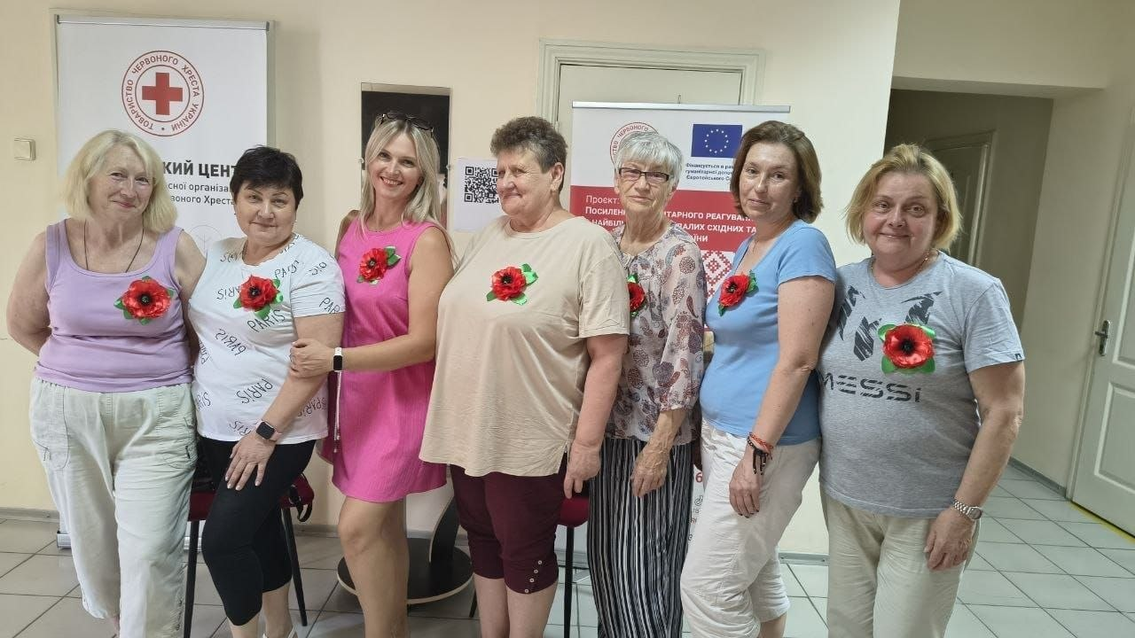 Група жінок різного віку стоїть у ряд у приміщенні організації, посміхається та позує для фото, маючи на одязі прикріплені червоні маки.