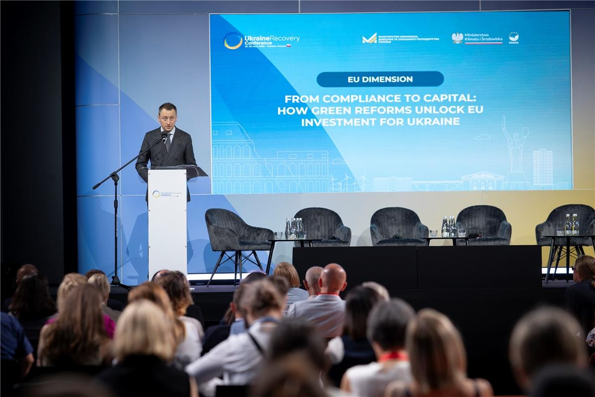 Чоловік у діловому костюмі виступає за трибуною на сцені перед аудиторією, на тлі великого синього екрана з англомовним текстом конференції та графічними силуетами міст.