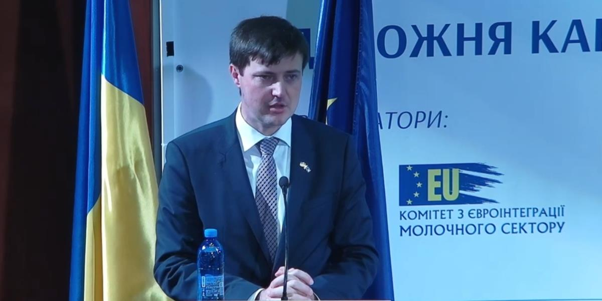Чоловік у діловому костюмі та краватці виступає за трибуною з мікрофоном на тлі державного прапора України та інформаційного банера з логотипом ЄС і написами українською мовою.