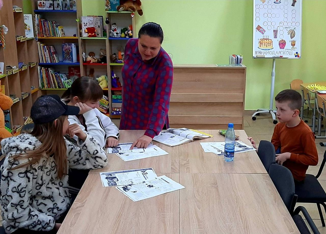 Жінка-викладач стоїть біля дерев'яного столу та показує матеріал у розгорнутій книзі дітям, які сидять навколо та слухають її в навчальній кімнаті з книжковими полицями.