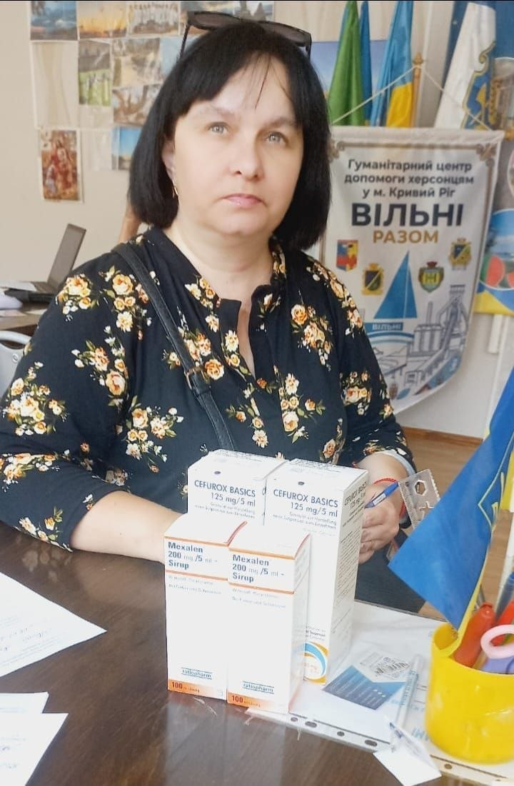 Жінка в темній блузці з квітковим принтом сидить за столом, на якому стоять коробки з медичними сиропами, на тлі банера гуманітарного центру та прапорів.