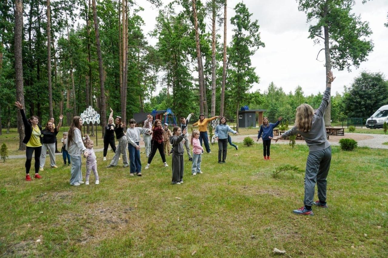 Група дітей та дорослих робить ранкову зарядку або повторює танцювальні рухи за аніматором на зеленій галявині серед дерев заміського комплексу чи табору.