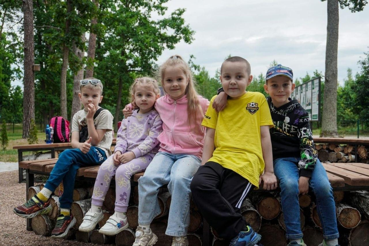 П'ятеро усміхнених дітей сидять поруч на дерев'яній лаві, облаштованій поверх складених колод, на тлі лісової зони або парку.