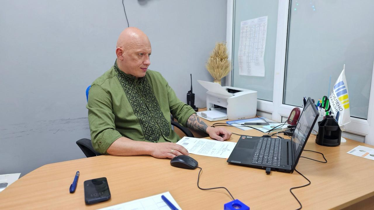 Чоловік у зеленій вишиванці сидить за робочим столом в офісі та дивиться на екран відкритого ноутбука. На столі перед ним лежать документи, комп'ютерна миша, смартфон, а поруч стоїть маленький прапорець із написом «Зроблено в Україні».