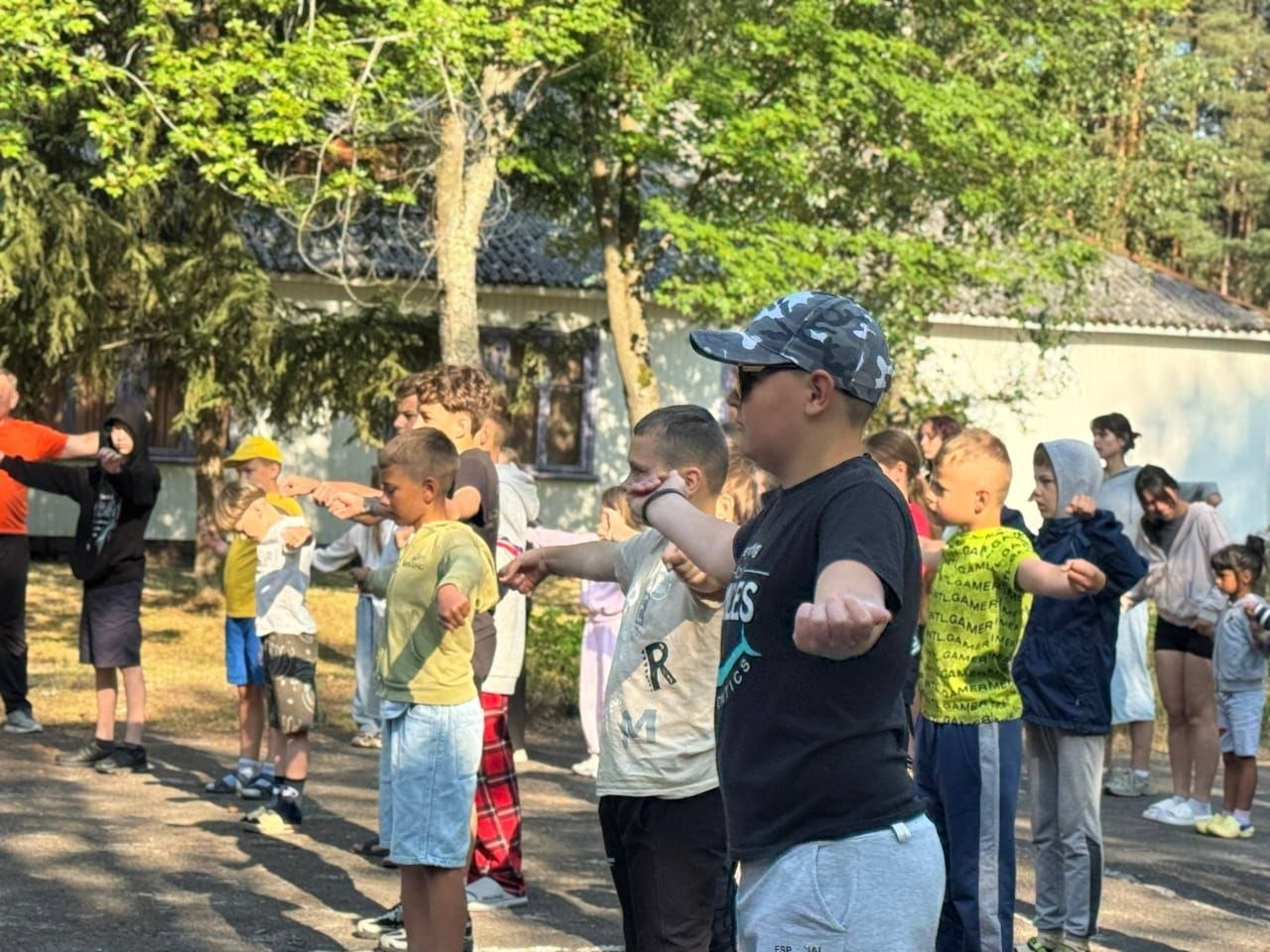Група дітей та підлітків виконує фізичні вправи на свіжому повітрі, витягнувши руки вперед, під час руханки чи тренування на території літнього табору на тлі дерев та одноповерхової будівлі.
